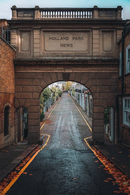 A narrow, cobbled street runs through an arched brick and stone gateway marked with a rectangular plaque inscribed 'HOLLAND PARK MEWS.' The archway, constructed from reddish-brown stone with detailed